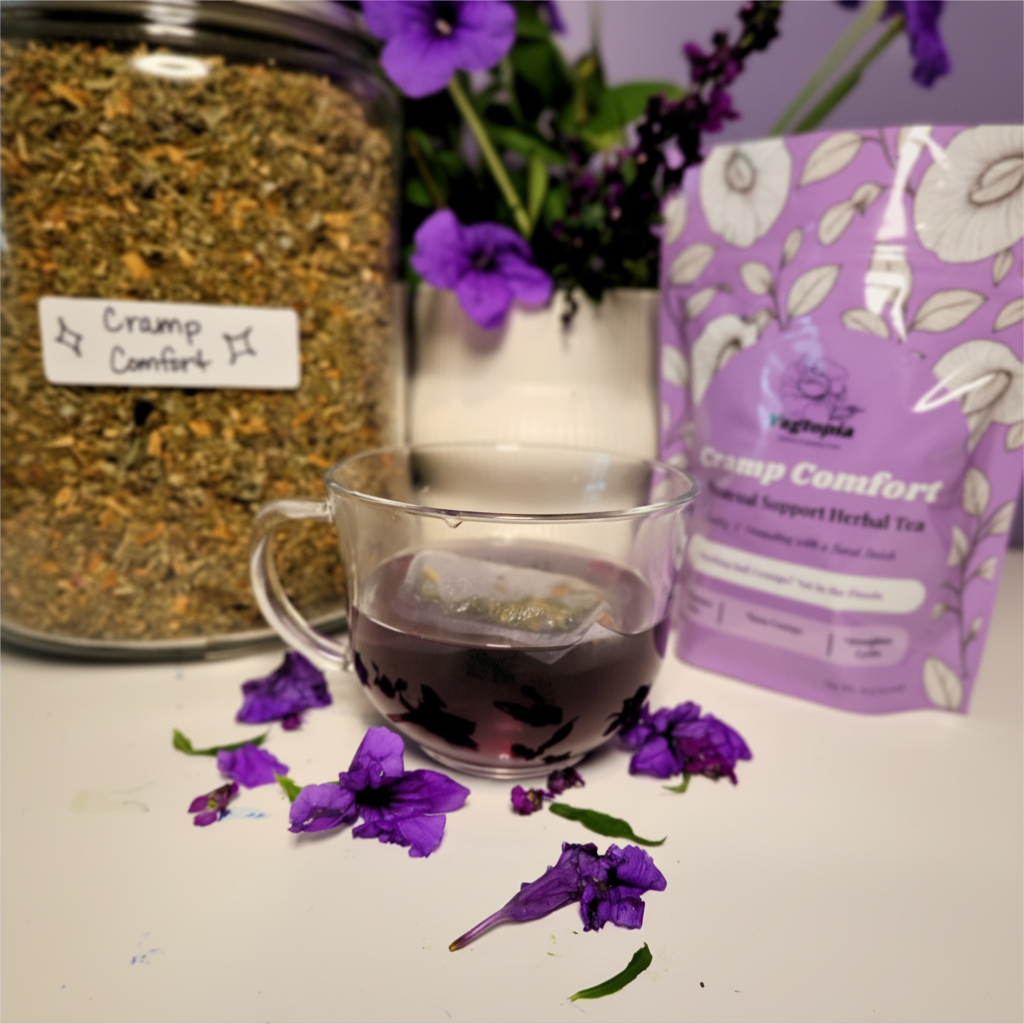 A clear glass mug filled with dark herbal tea sits on a white surface, with a branded pouch of Vagtopia Cramp Comfort herbal tea to the right and a large jar of loose herbal material to the left. Purple flowers and petals are scattered around the mug.